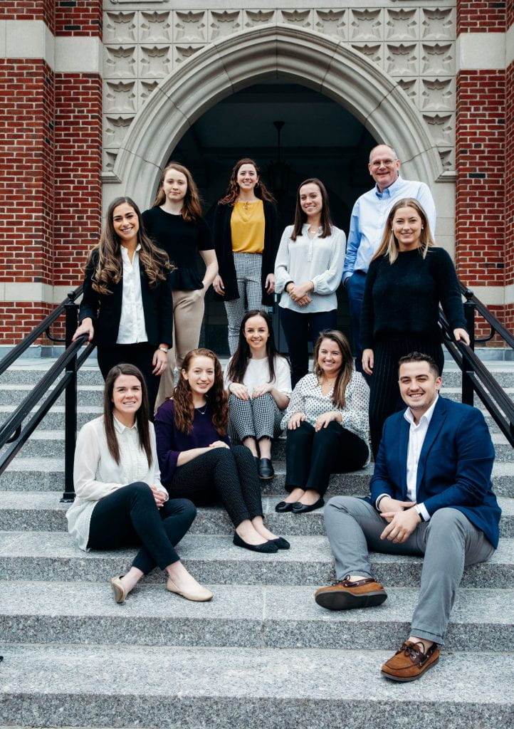 Dr. Robert Hackey, professor of health policy and management, standing at right, joins several of the students who are writing a volume in a book series on healthcare with Hackey and Dr. Todd Olszewski, associate professor of health policy and management. Students are, sitting from left, Rose Shelley ’19, Delaney Mayette ’20, Erin Walsh ’20, Amanda McGrath ’20, and Keith Vieira ’21, and standing, Nicolette Greco ’20, Theresa Durkee ’20, Anne Capozzoli ’20, Shannon McGonagle ’19, and Morgan Bjarno ’19.