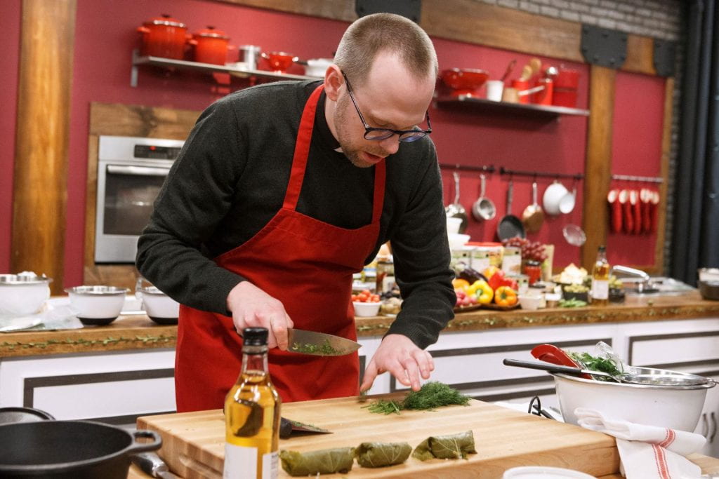 Father Adam, wearing an apron, chops herbs on the Worst Cooks set.