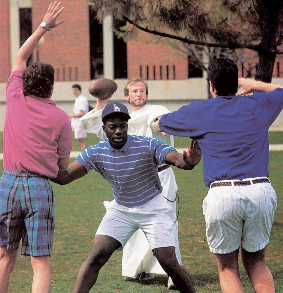Father Shanley plays quarterback on the Slavin Center lawn during his time as residence hall director and DWC instructor. HIs blocker is Earl Smith '92.