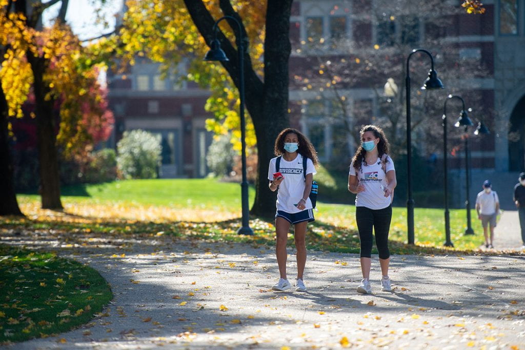Two women, wearing masks, walk across campus on a warm November day.