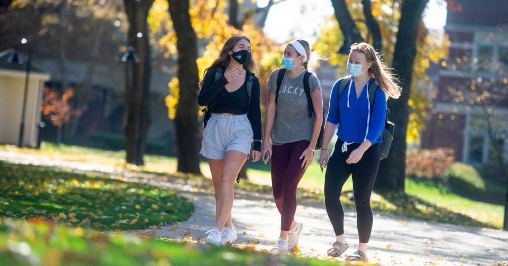 Three women wearing masks walk on a path at Providence College in autumn.