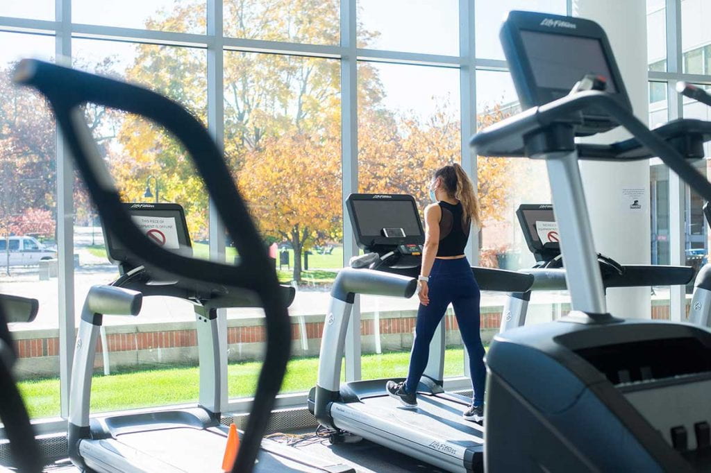 A female student works out on a treadmill in the Concannon Fitness Center facing windows showing an autumn scene on campus.