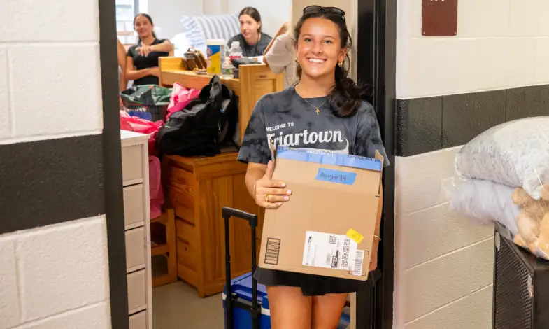 Class of 2029 student holding a box and smiling in front of her dorm room during Move-In Day