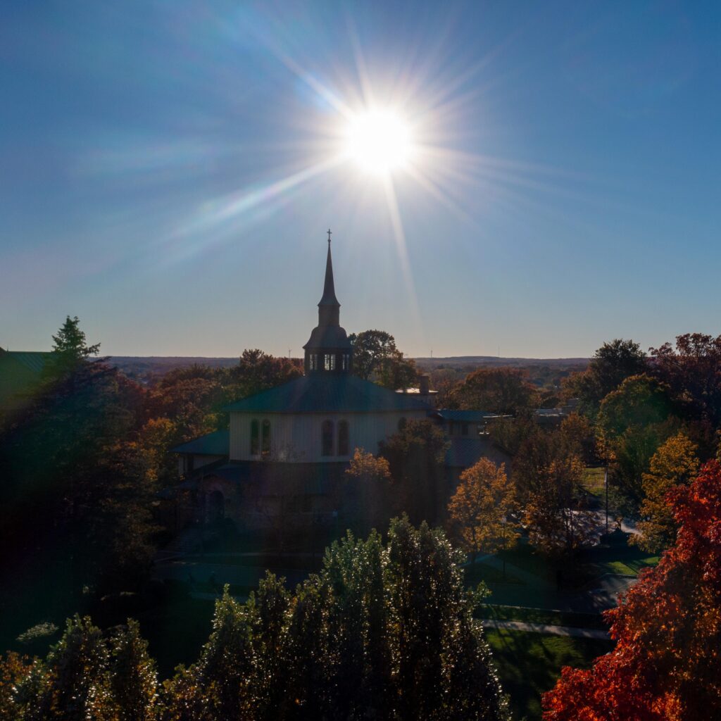 Drone shot of St. Dominic Chapel, with sun flare in the sky and surrounded by fall foliage