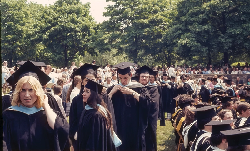 Undergraduates in line for commencement with green trees and spectators.