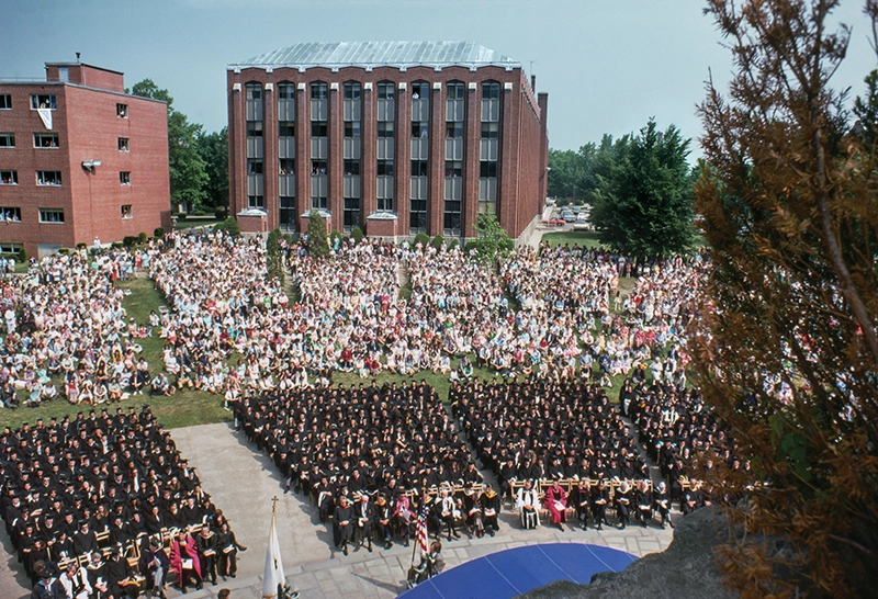 An aerial view of the 1975 commencement taken from the War Memorial Grotto, looking toward McDermott Hall, left, and Aquinas Hall, right. Graduates in charis and spectators behind them on the lawn.