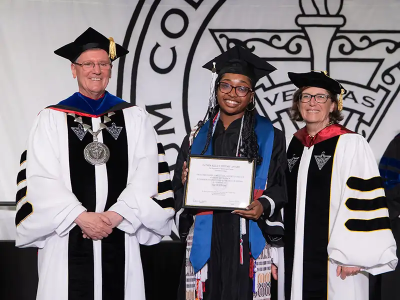 Angie Pierre '25, dressed in her graduation cap and gown and wearing numerous honor cords and stoles, receives the Father Reilly Award for history scholarship at the Academic Awards Ceremony. She is standing with College President Father Sicard and Sylvia Maxfield, the interim provost.