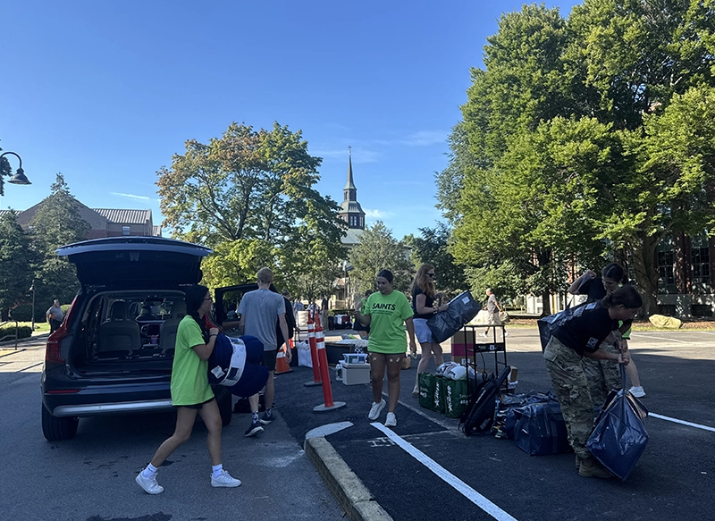 Students in yellow T-shirts unload belongings from an SUV and place them on the sidewalk.