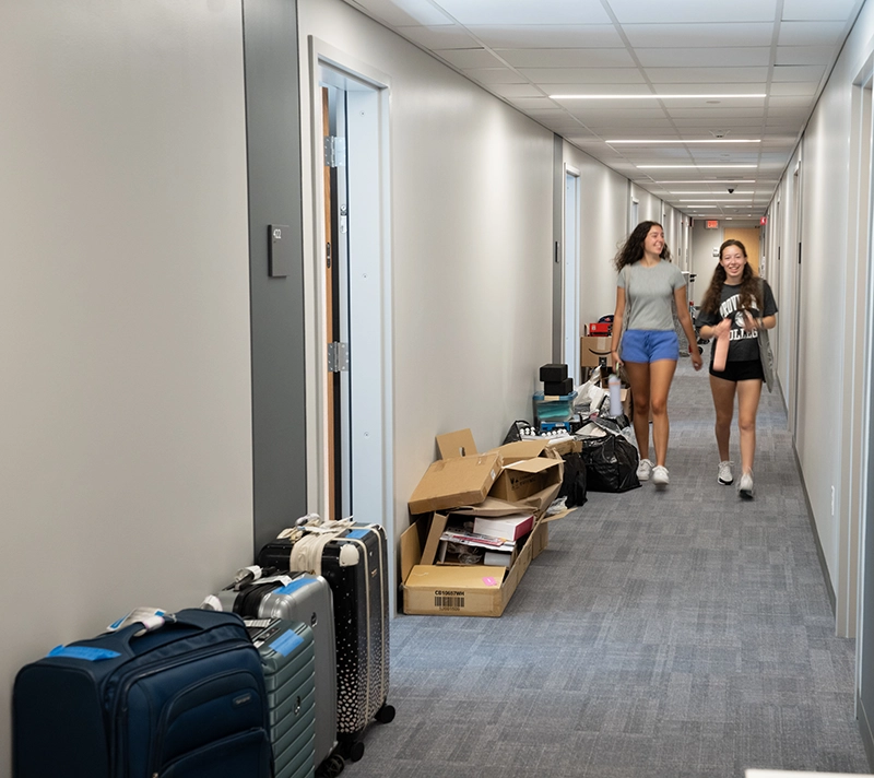 Two young women in a carpeted hallway with suitcases along the sides
