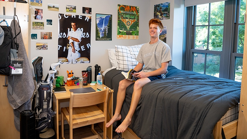 Young man seated on a bed next to windows, posters cover walls, desk nearby