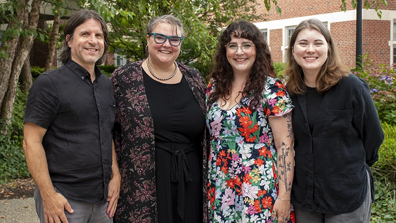 Members of the digital team from Phillips Memorial Library, one man and three women, stand outside in front of a tree on campus.
