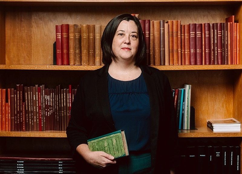 A woman holding a book with a green cover from the 1930s stands in front of a library shelf of books.