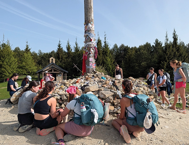 Women kneel before the base of a wooden cross. The cross is surrounded by mementos left by pilgrims on the way to the cathedral in Spain.
