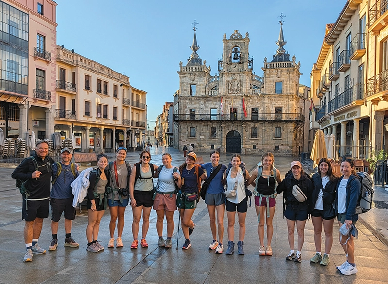 Two men and 11 women who are student athletes stand in a plaza in Spain with a cathedral behind them.