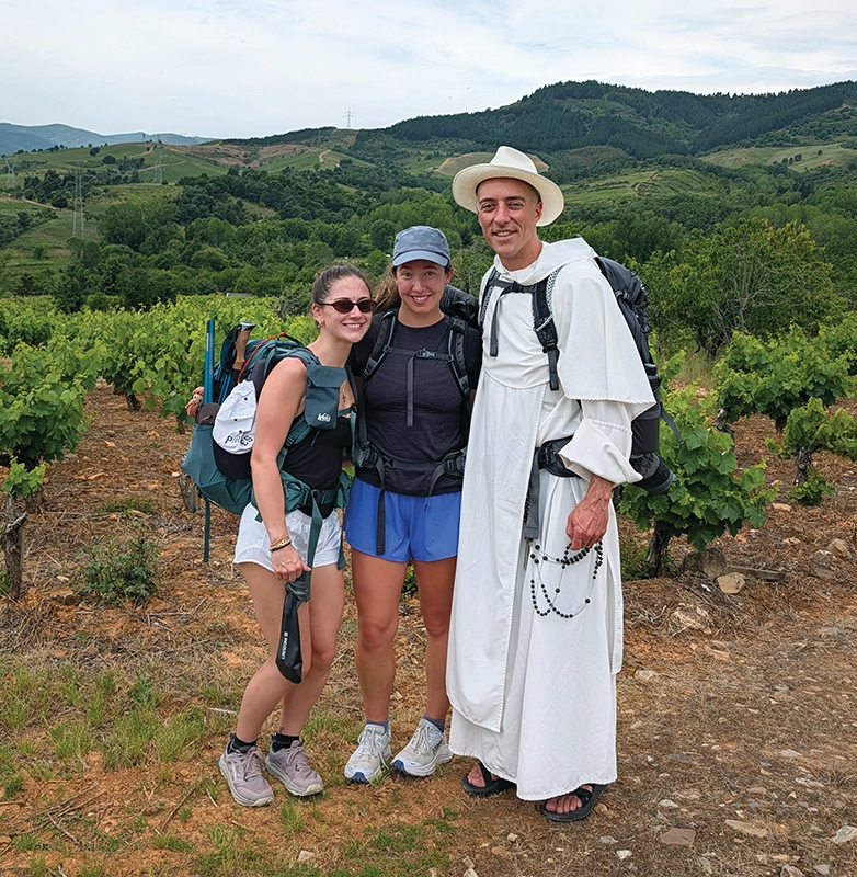 Two students in hiking gear pose with a priest in white robes along a trail with mountains behind them.