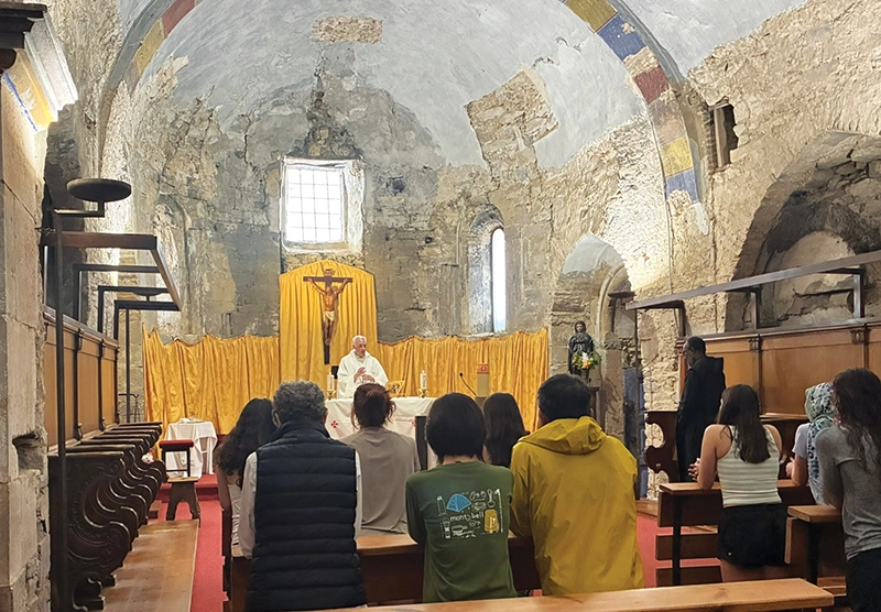 A priest celebrates Mass in a church in Spain with walls lined with stone as students kneel in prayer.