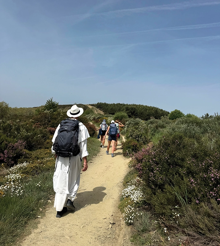 A priest in a white robe, hat, and backpack follows students along a curving dirt trail lined with green bushes and white flowers.