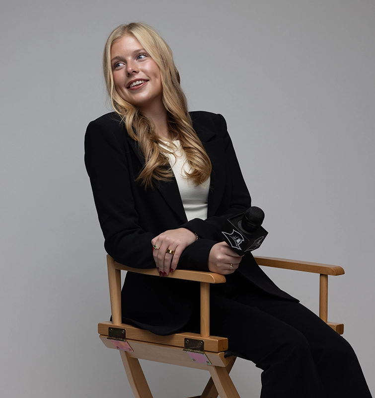 Caroline Austin '26 sits in a director's chair while holding a microphone with the Friar head logo.