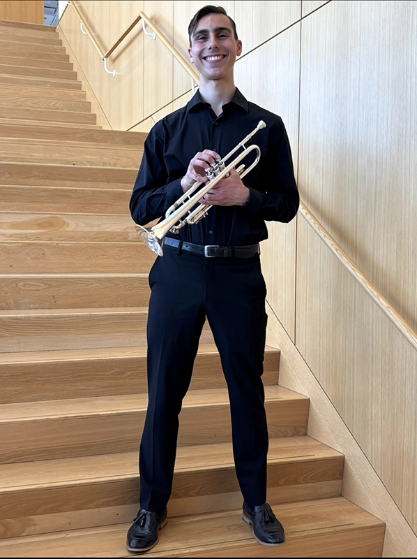 Nick Cassarino '27 holds a trumpet as he stands on a staircase, dressed in black shirt and pants before a performance.