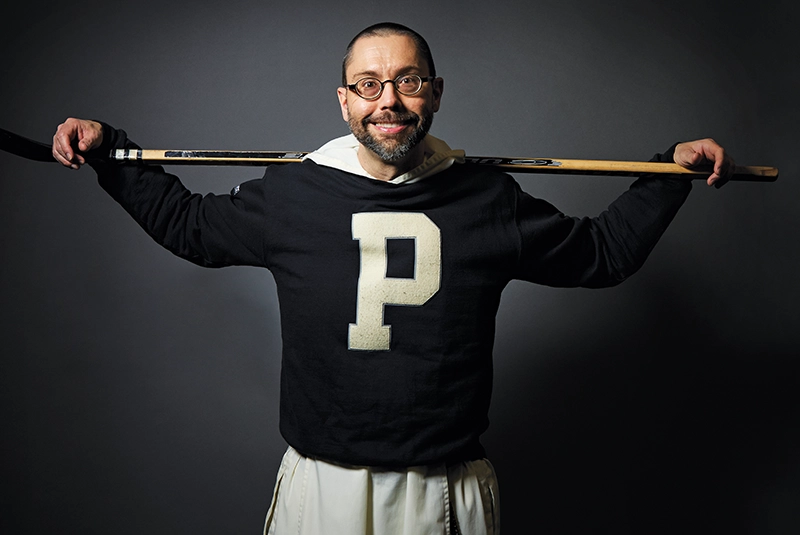 A priest wearing a black sweater with a white "P" insignia poses with a hockey stick.