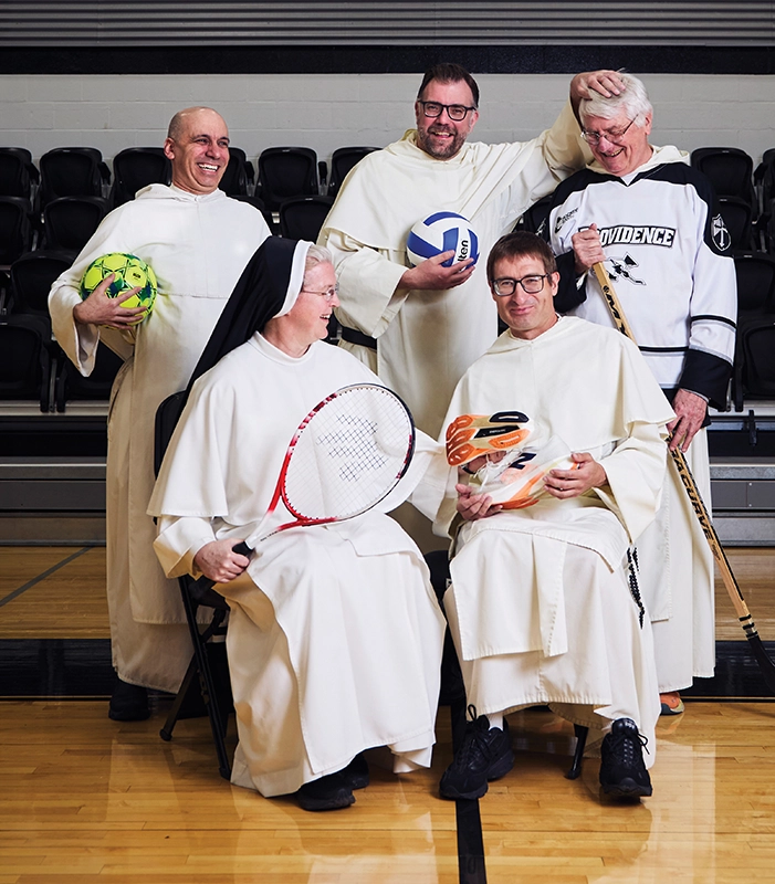 Three priests and a religious sister, all holding sports equipment, laugh during a photo session in a college gym.