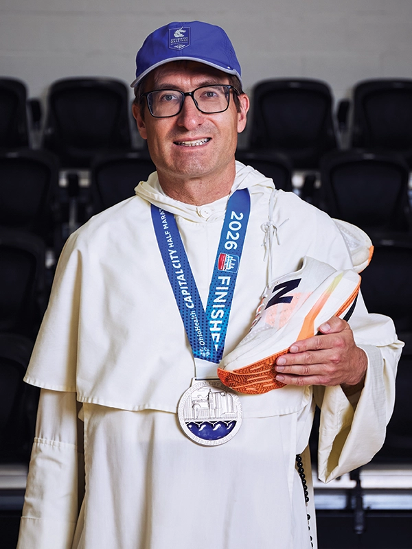 A priest in a white robe holds a running shoe. Around his neck is a medal showing that he finished the 2026 Capital City Half Marathon.