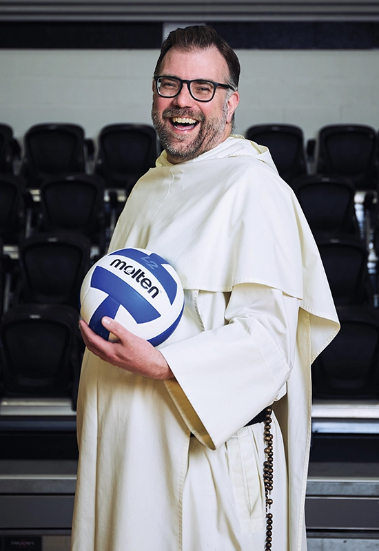 A priest wearing a white robe laughs while posing with a volleyball.
