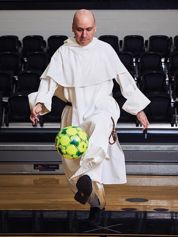 A priest in a white robe juggles a soccer ball in a gym.