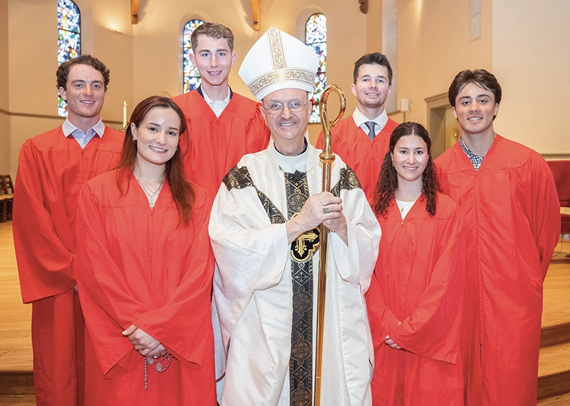 Six student-athletes, men and women, dressed in red robes, pose in the chapel with the bishop of the Diocese of Providence.