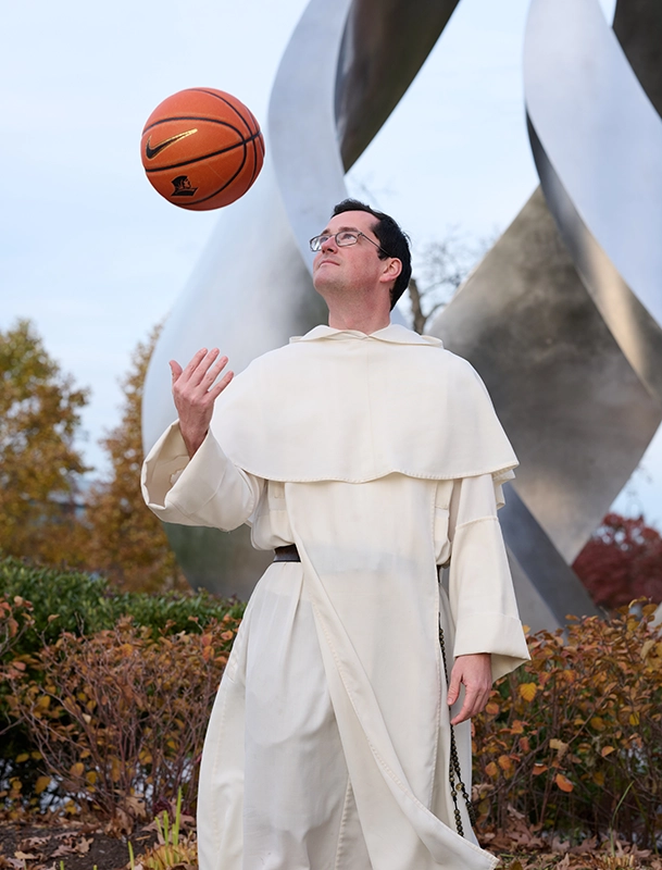 A priest in a white robe tosses a basketball in the air. Behind him is a metal sculpture representing a flame.