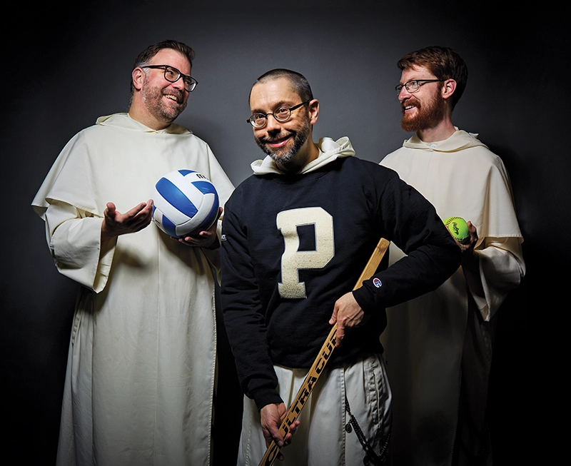 Two priests in white robes hold a volleyball and a softball. A priest in the foreground, wearing a black sweater with a "P" insignia, holds an ice hockey stick.