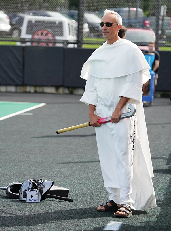 Priest in white robe on side of field hockey game holding a field hockey stick.