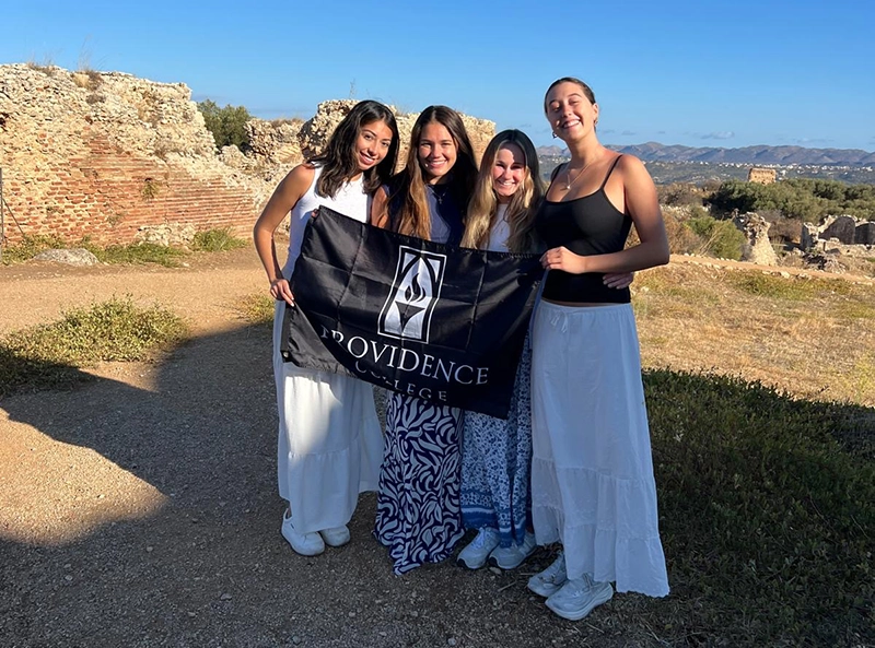 Four women holding a banner reading "Providence College" stand on a grassy point overlooking ruins and mountains in Crete.