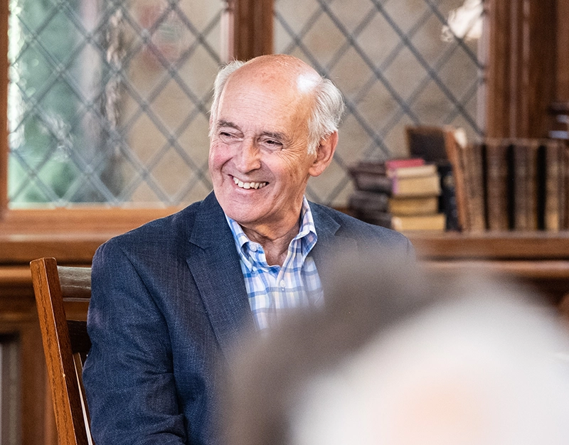 Joe Brum smiles in appreciation as he listens to a story during his class reunion. Piles of books behind him.