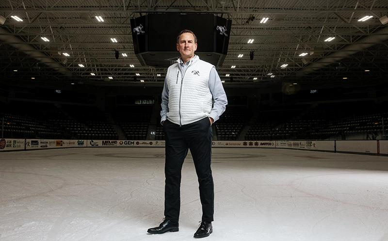 Joe Hulbig, wearing a gray Friar vest with hands in his pockets, stands on the ice on the Providence College hockey rink.