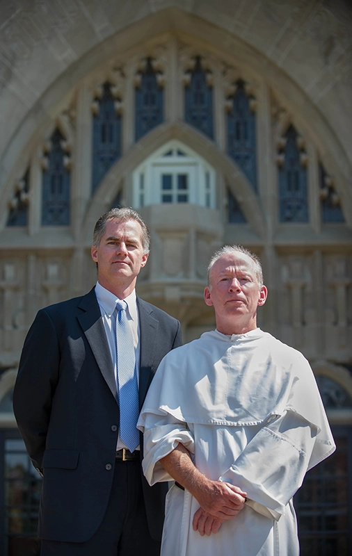 Two men, one a priest wearing a white habit, stand together outside a college building.