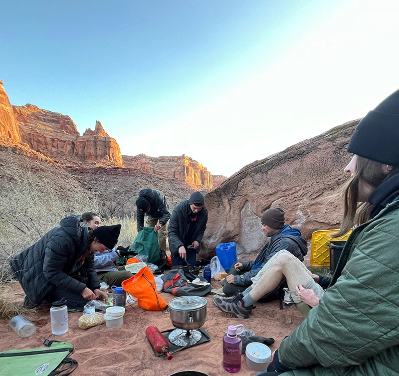 Students wearing beanies sit on the ground preparing a meal with mountains in the background.