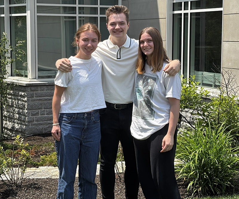 Three students, two women and one man, outside the college's science building in the summer, surrounded by green plants.