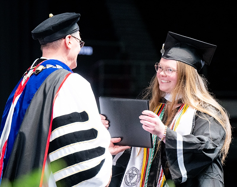 Female student wearing cap and gown accepts diploma from the college president, who is in full academic attire.
