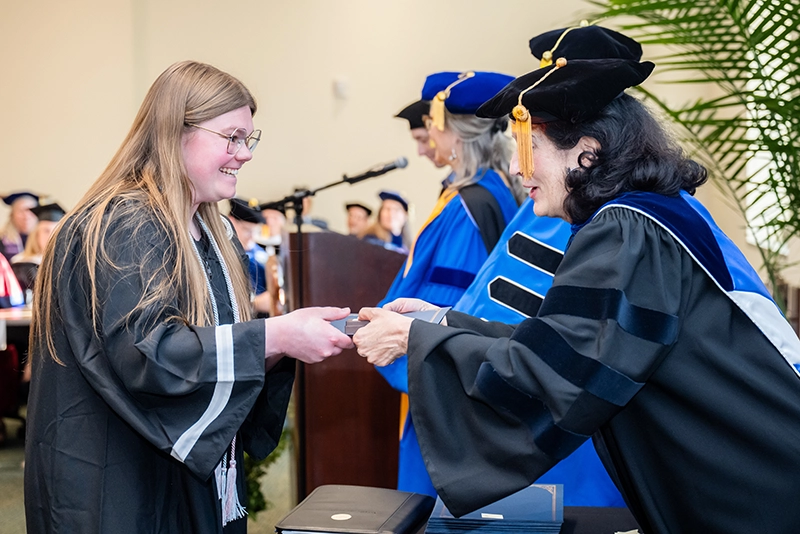Female student wearing graduation robe receives certificate from female professor in full academic attire.