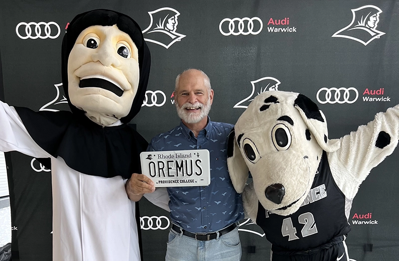 A man holds a license plate accompanied by the college mascots, a Dominican friar and a Dalmatian.
