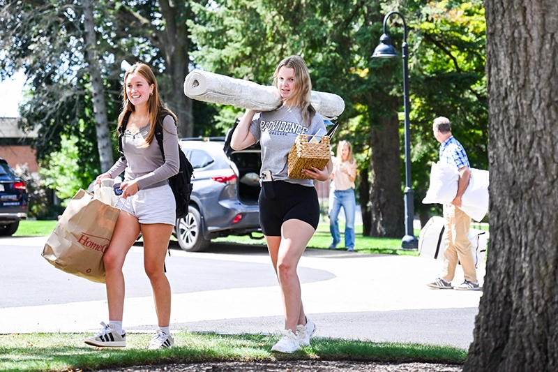 Two young women carry belongings -- a rug and in a shopping bag -- across campus on Move-In Day.