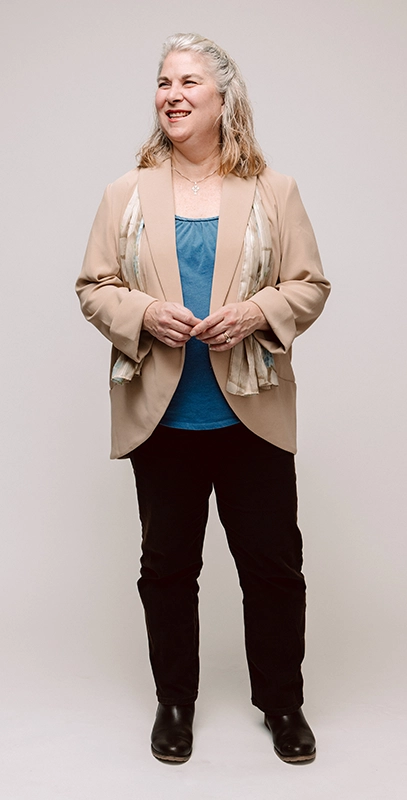 Sharon Ann Murphy stands with hands touching wearing a tan jacket and black pants.