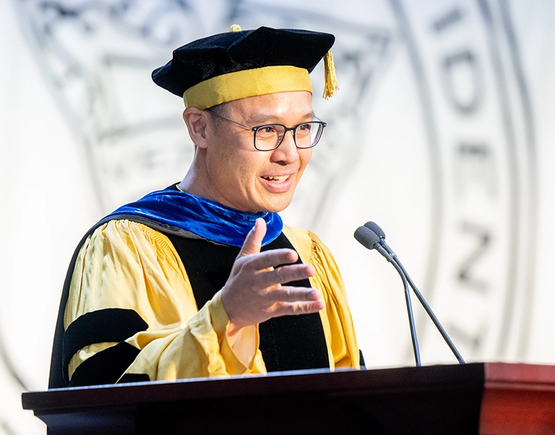 Man in yellow and black cap and gown at lectern