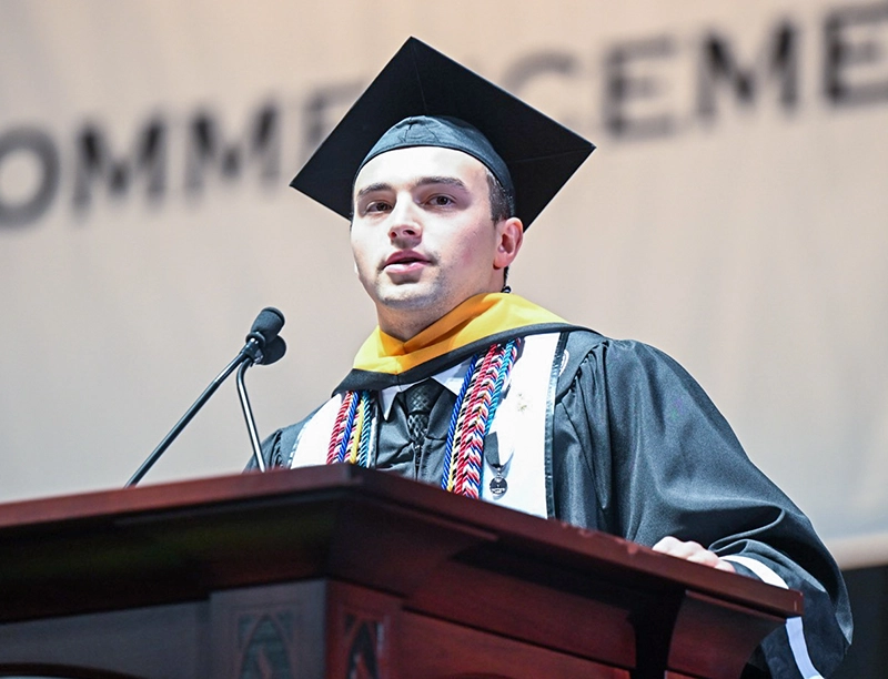 Young man in cap and gown at lectern with microphone.