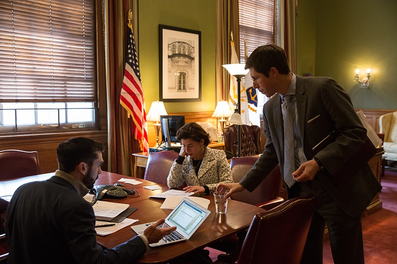 Mike Raia gestures at a laptop, RI Governor Gina Raimondo sits behind a desk, Providence Mayor Brett Smiley stands nearby.