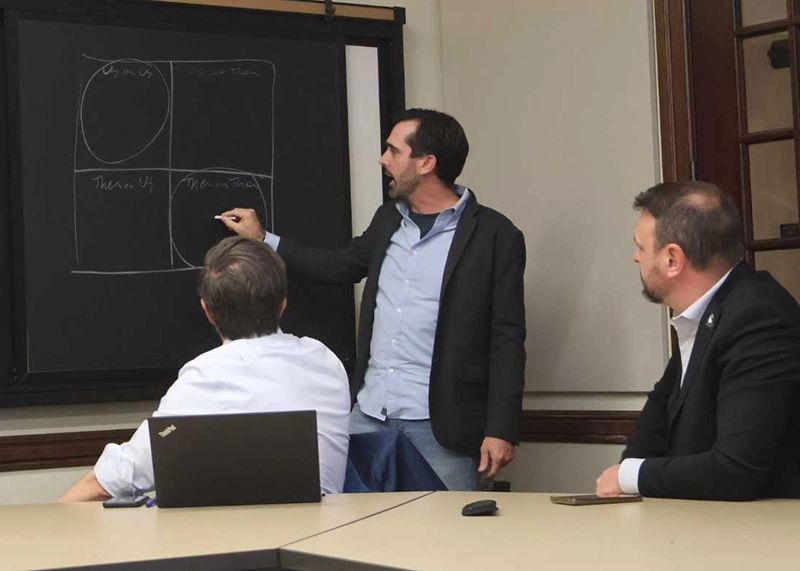 Mike Raia draws on a chalkboard as two professors sit and listen to him.