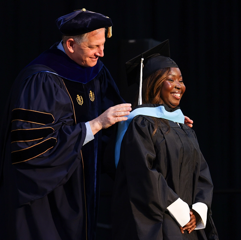A professor wearing academic regalia places a blue hood around the shoulders of a female student.