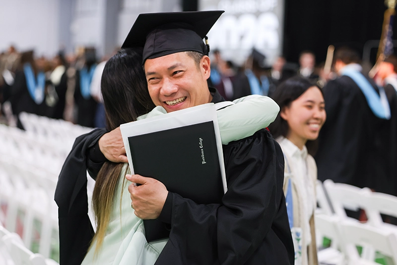 A student in a cap and gown holding a diploma is hugged by a woman.