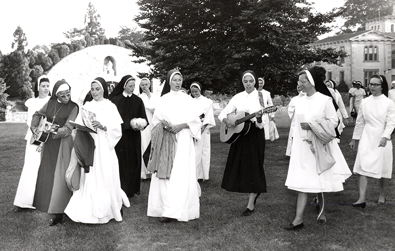A group of religious sisters in white habits walk across the campus lawn. Two are strumming guitars.